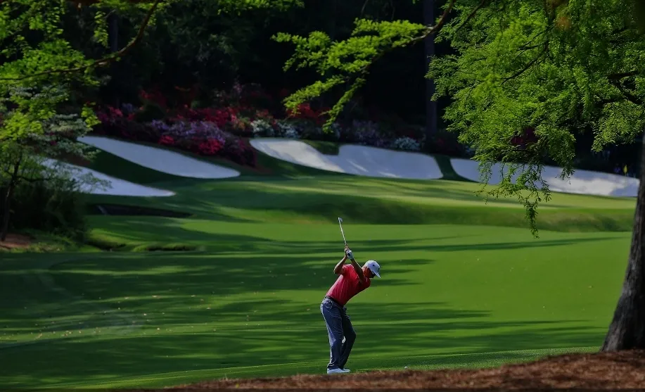 A golfer playing a shot beside a Masters green bordered by white bunkers, trees and flowering shrubs.